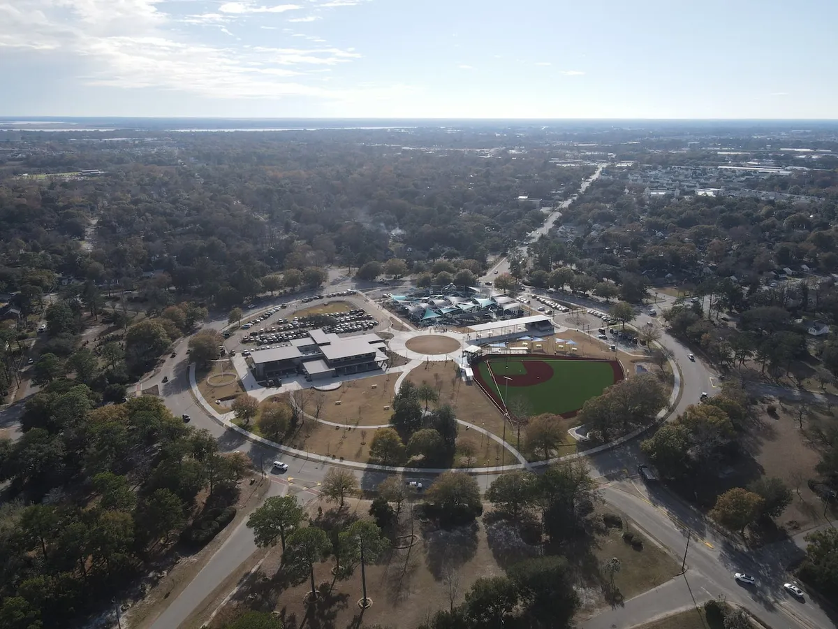 A broad exterior shot showing a community baseball field adjacent to a community center, with a busy parking lot and lush park surroundings. The image conveys the energetic atmosphere of the neighborhood, making it ideal for outdoor breaks or evening walks.