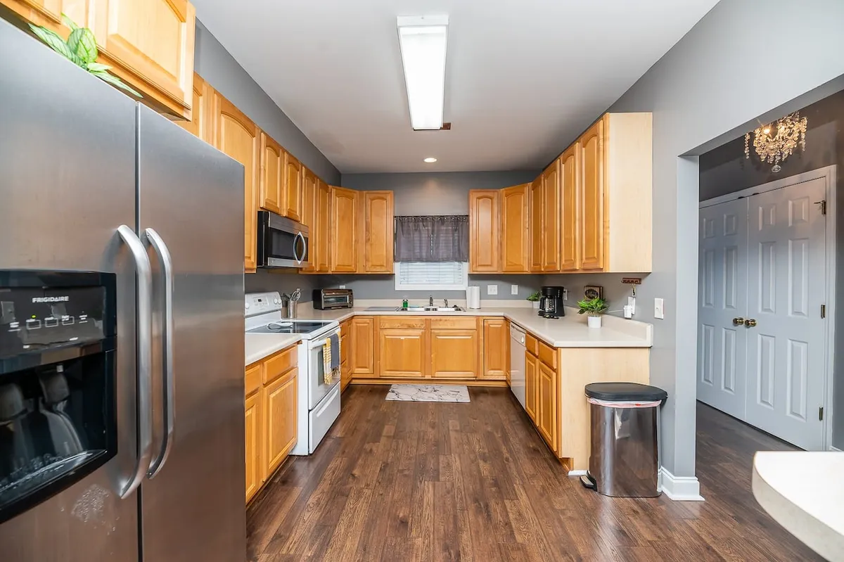 This image displays a modern kitchen with warm wooden cabinetry, gleaming stainless steel appliances, a spacious kitchen island, and sunlight streaming in through a window. The scene conveys a lively, functional space suitable for both cooking and gathering.