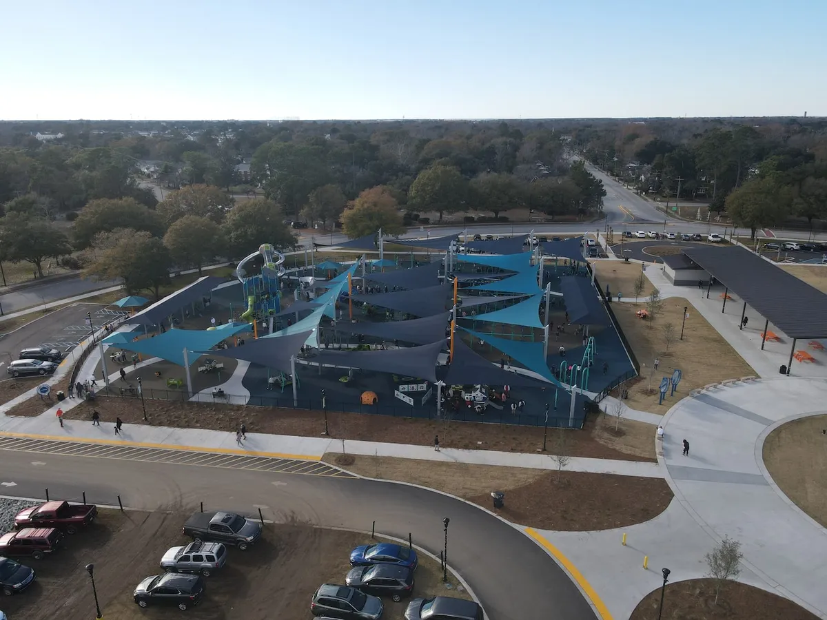 An exterior view of a vibrant community park featuring modern play structures under shade sails, children engaging in play, walking paths, and a visible parking area. The scene captures a lively, welcoming neighborhood setting ideal for walks, quick errands, or relaxation after work.