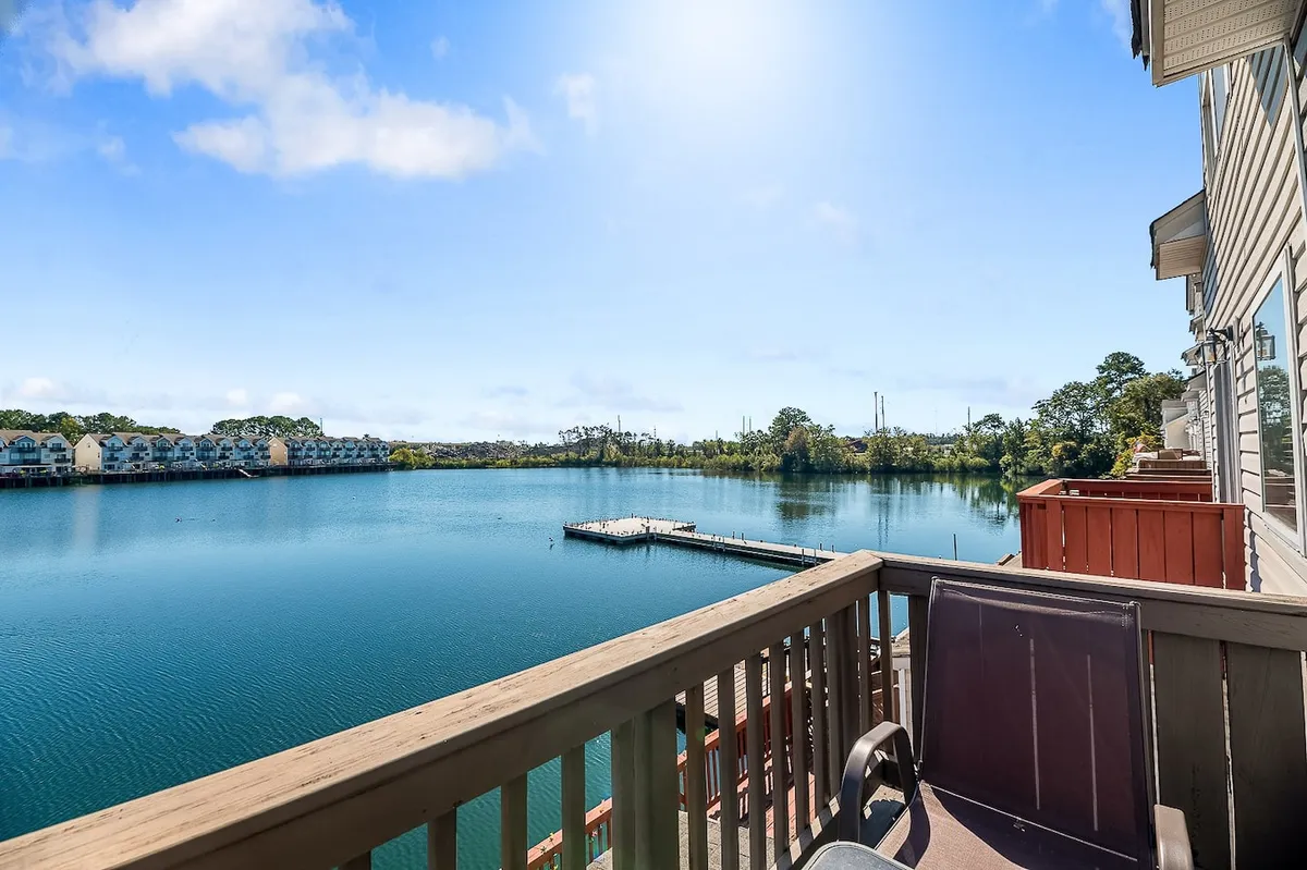 A serene view of calm water with a pier, neighboring houses, and a balcony railing. The setting exudes peacefulness and hints at potential for fishing, boating, or simply enjoying the waterfront atmosphere with family.