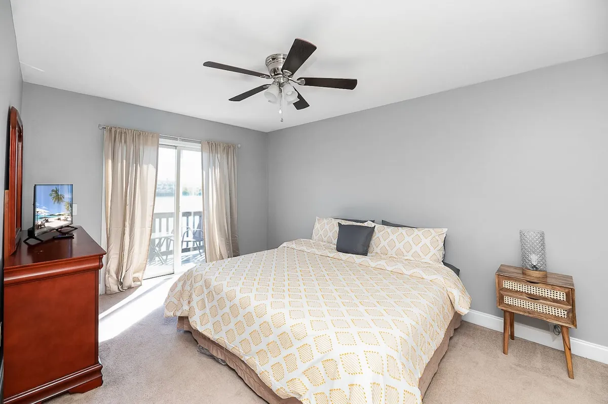 A tranquil bedroom with a king-size bed, flat-screen TV, wooden dresser, and nightstand. Sunlight streams in through the curtained window, setting a peaceful tone for relaxation or a cozy movie night.