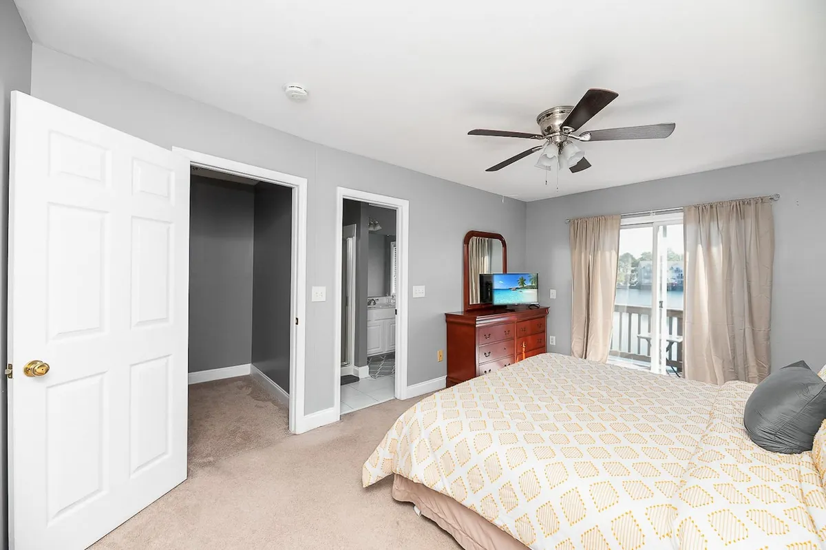 A cozy bedroom featuring a bed with patterned bedding, a wooden dresser, and a window with soft curtains letting in natural light. The space feels intimate and quiet, perfect for restful nights after a busy day.