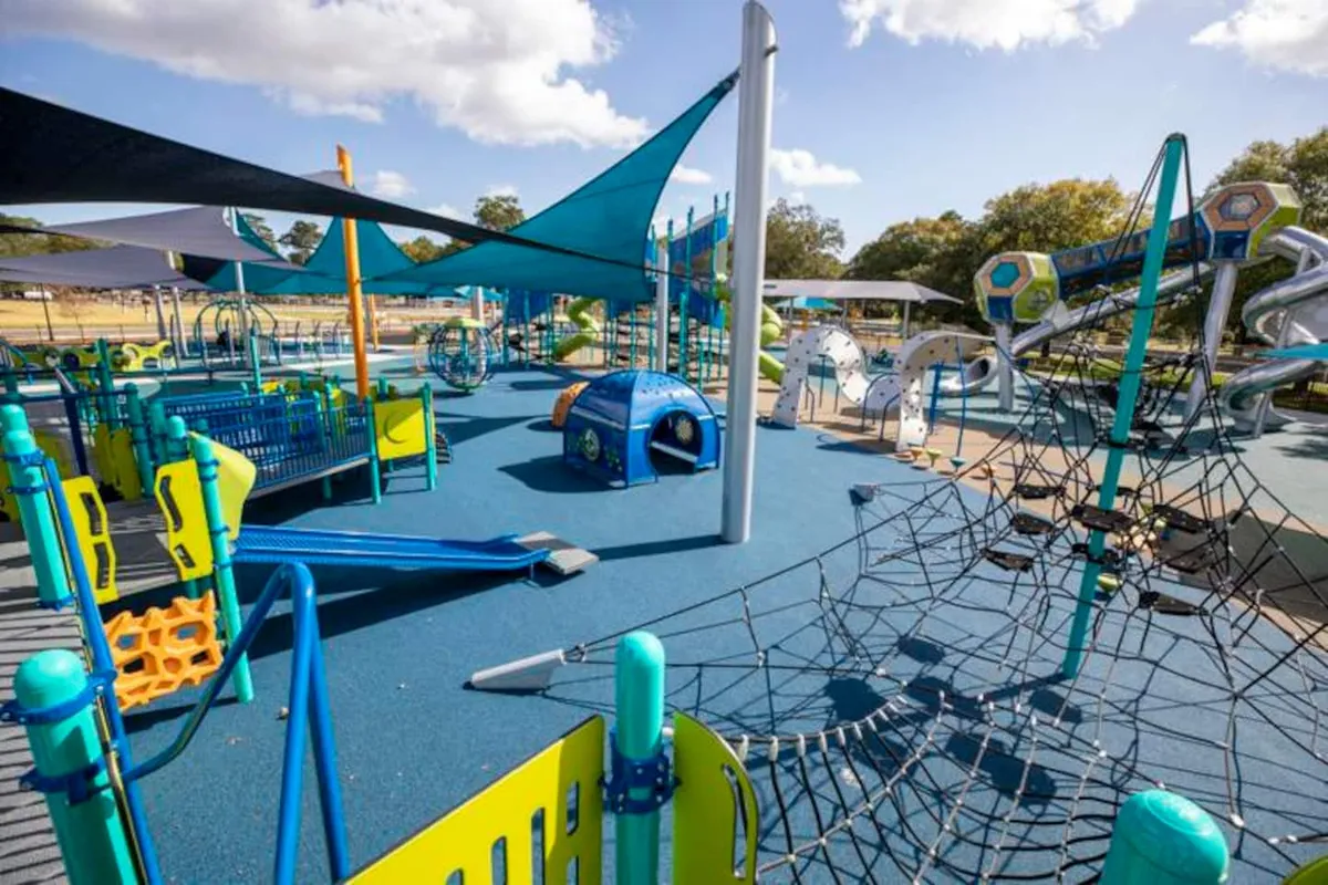 The image captures a vibrant playground scene with a climbing net, slide, multiple play structures, tunnels, and shade sails. Children are seen playing and families are gathered, enjoying a lively outdoor environment perfect for family outings and gatherings.