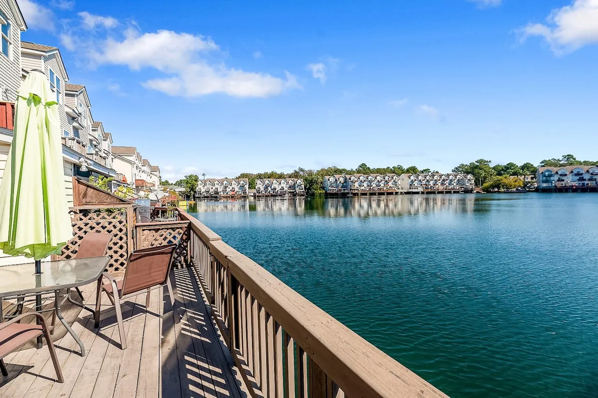 A wooden back deck overlooking the calm lake, furnished with outdoor seating and colorful umbrellas. The inviting setup is perfect for enjoying coffee in the morning, gathering with friends, or simply relaxing while taking in the scenic water views.