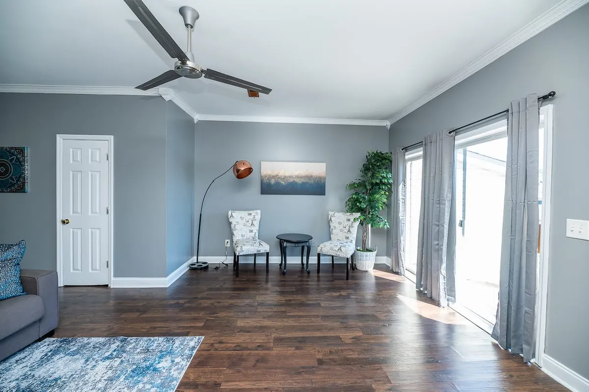 A sun-filled living room featuring a plush sofa and armchairs arranged for conversation, accented by tasteful artwork and a leafy plant. The space is open and inviting, with ample natural light streaming through large windows, highlighting its modern, airy ambiance.